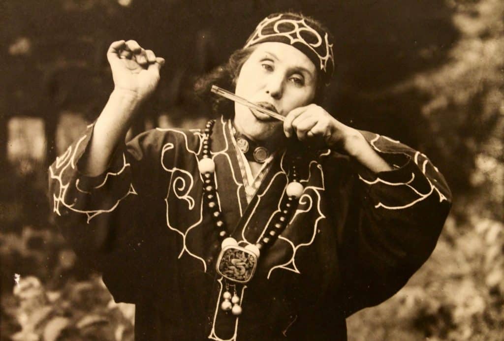 old photo of Ainu woman playing a wooden instrument in traditional clothing in mid pose