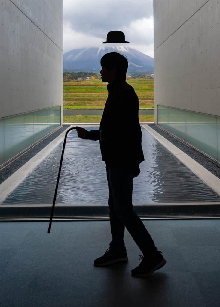 man posing in front of Mt. Daisen with hovering hat and cane at the Shoji Ueda Museum of Photography