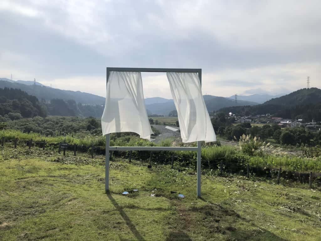 A so-called window in the middle of Japanese countryside, with white curtains moving with the wind. Artwork