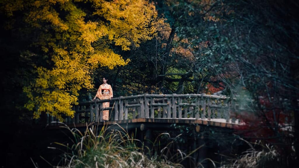 A girl wearing a kimono stands on a bridge surrounded by trees in Japan
