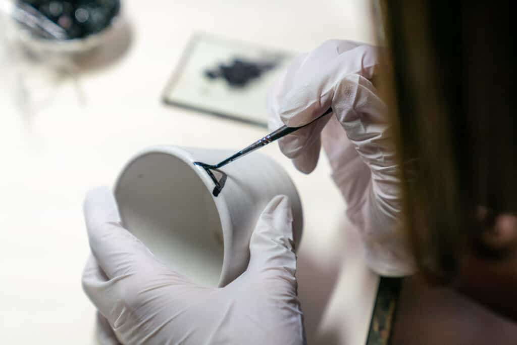 Person adding black lacquer to a white ceramic cup, performing the Japanese art of kintsugi