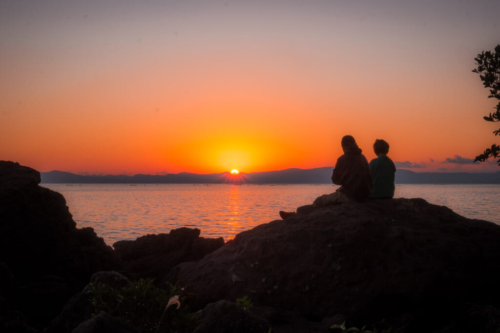 couple sitting on large rocks watch sunset at Sugawara Shrine in Kagoshima, JApan