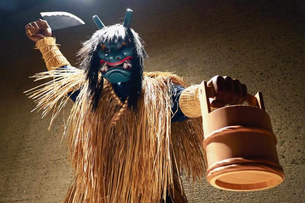 blue masked namahage figure in straw costume with knife and pail