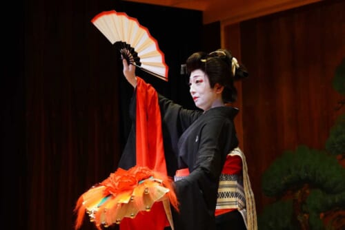 Woman in dark kimono with white make up on a stage holding a fan