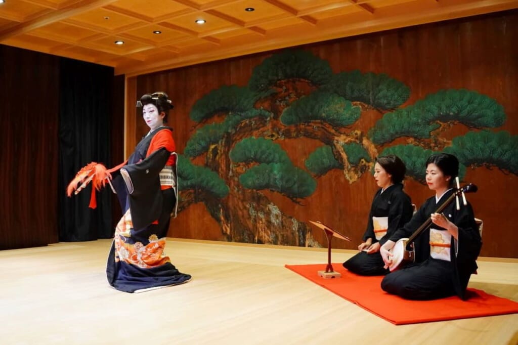 Three women in dark kimono on a stage, two kneeling and one standing