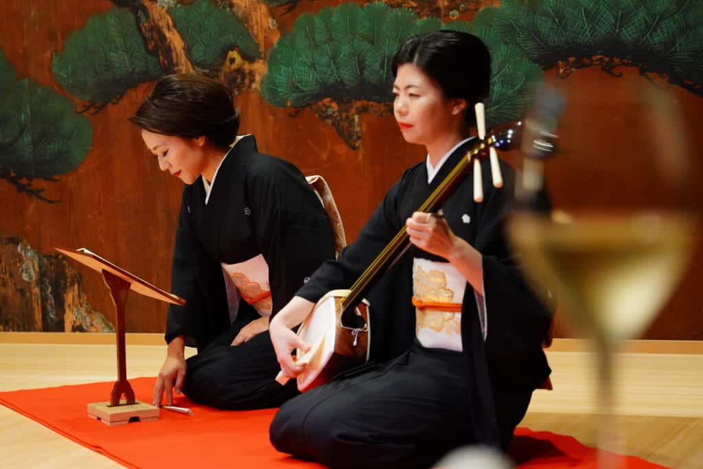 Two women kneeling in dark kimono on a stage with hair tied up, one holding a shamisen stringed instrument. 