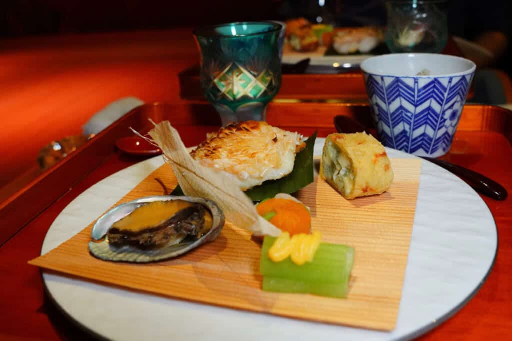 A white plate with a bamboo mat and various Japanese food, including fish