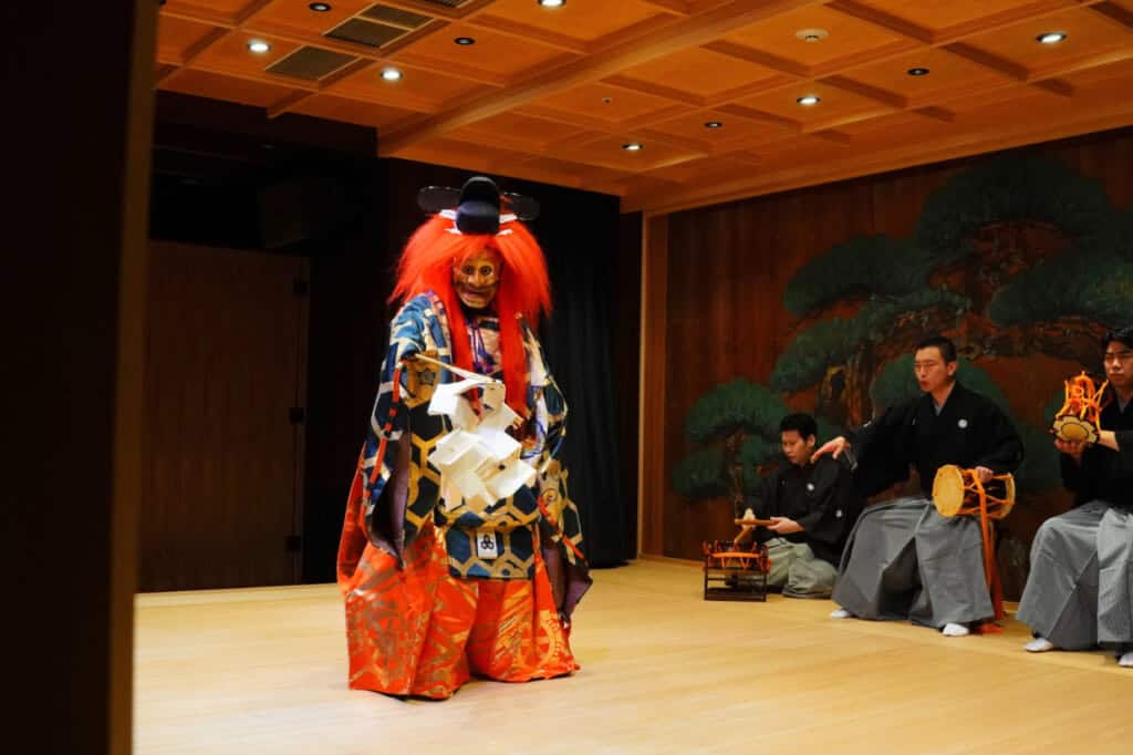 Three instrumentalists seated and a Noh-mask wearer standing on a stage