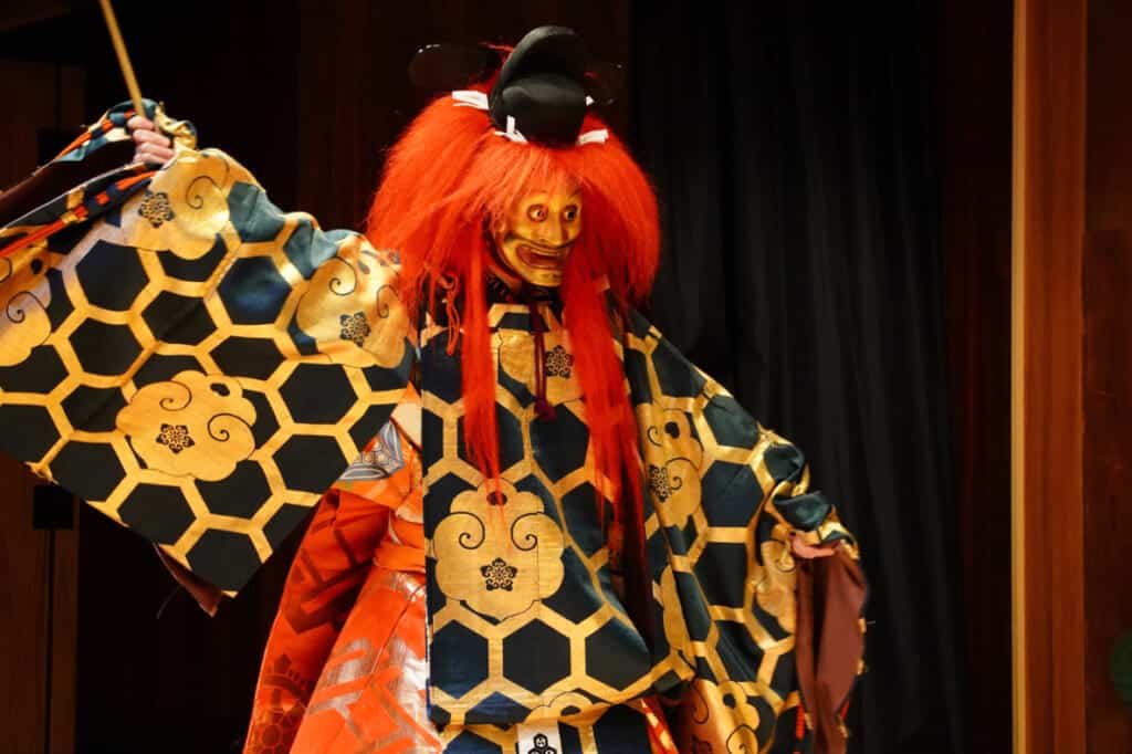 A Noh performer with bright red, long hair, a golden mask, and navy and gold clothes