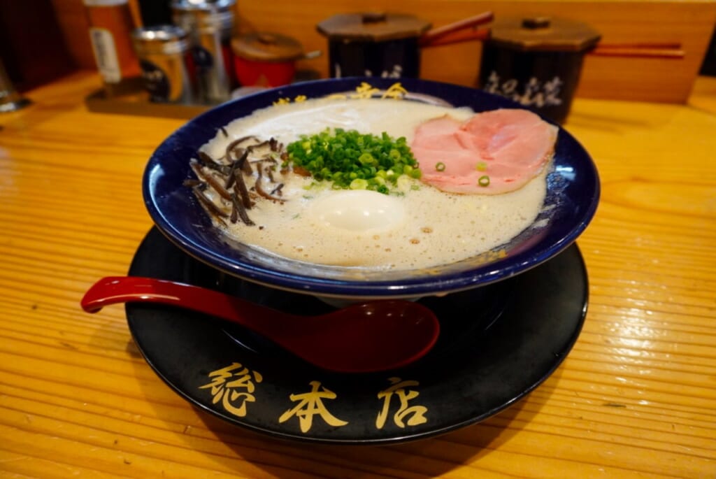 A blue bowl of frothy and creamy tonkotsu ramen with various toppings on a wooden table.