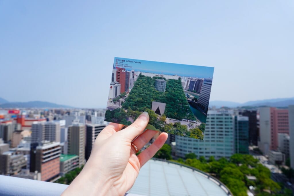 A view of Fukuoka city from above with a woman's hand holding a postcard of a green building in front of it.