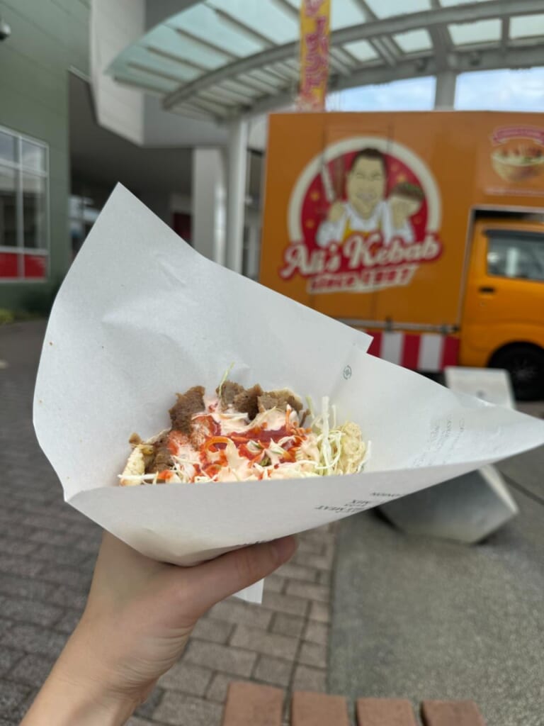 A woman's hand holding a pita kebab sandwich in front of an orange kebab truck