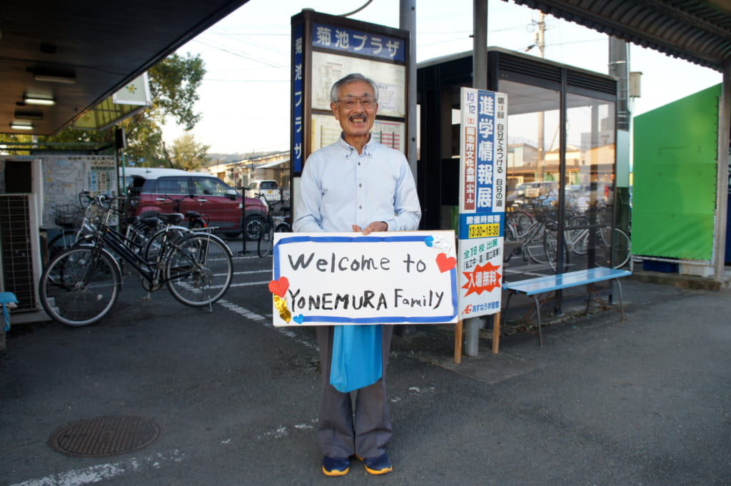 M. Yonemura, deant la gare routière avec une pancarte "welcome to Yonemura family"