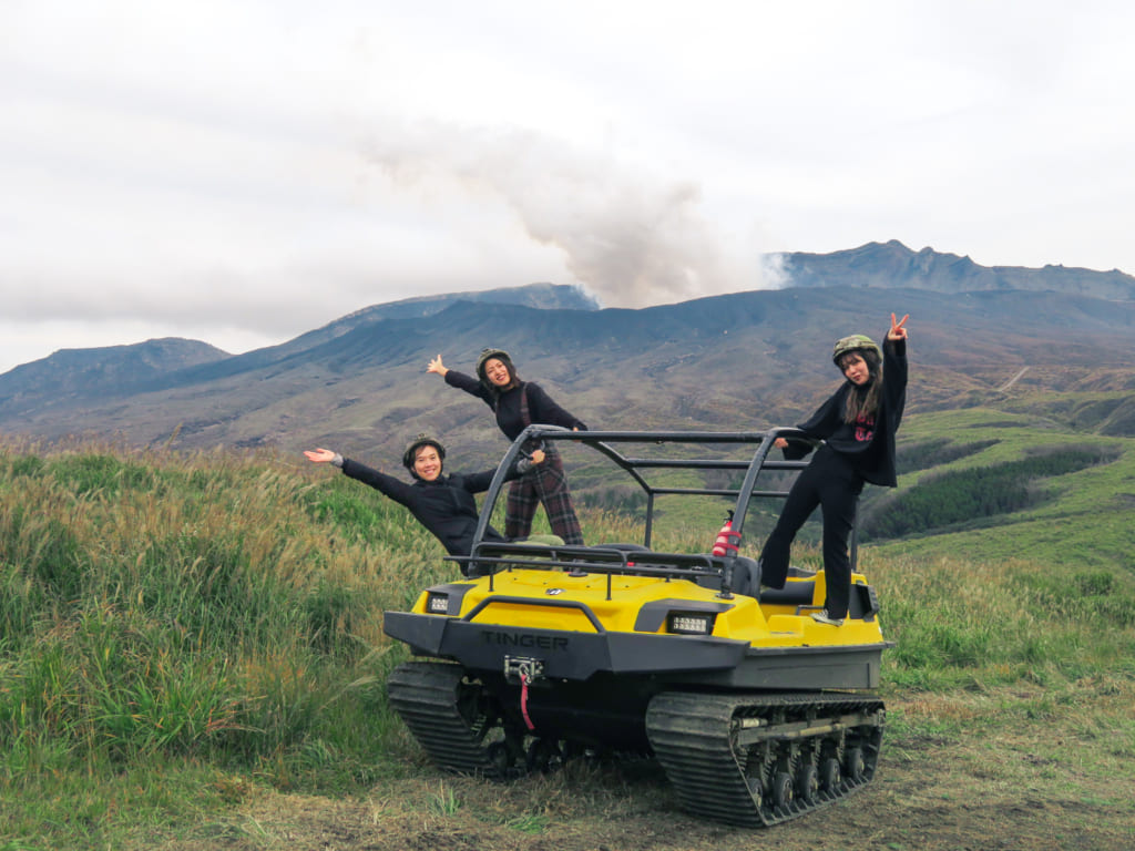 Mika accompagnée de deux autres personnes posent sur le camion tout terrain devant une éruption du mont Aso : une des activités de plein air à pratiquer à Kumamoto