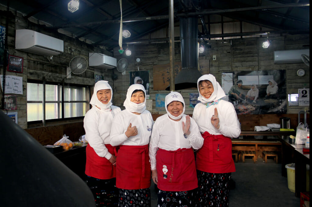 In a wide room, 4 women look at the camera with big smiles. They wear white shirts and hats, dark skirts and red aprons.