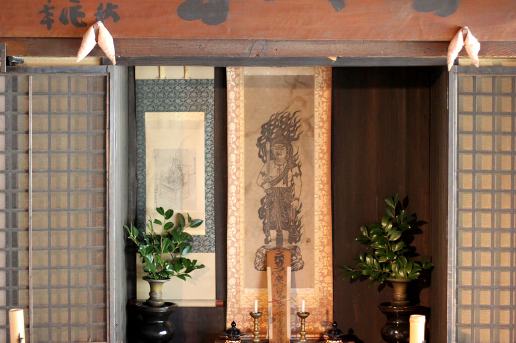 Des portes ajourées en bois ouvertes sur un dessin à l'encre représentant une figure divine. Des bougies et des vases sont posés de chaque coté.
