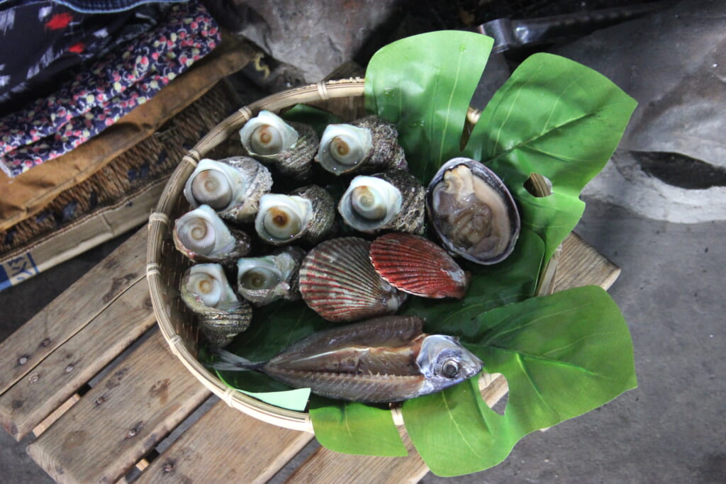 Un plateau de fruits de mer est posé sur un tabouret en bois. On y voit differents coquillages et un poisson posés sur une large feuille verte.