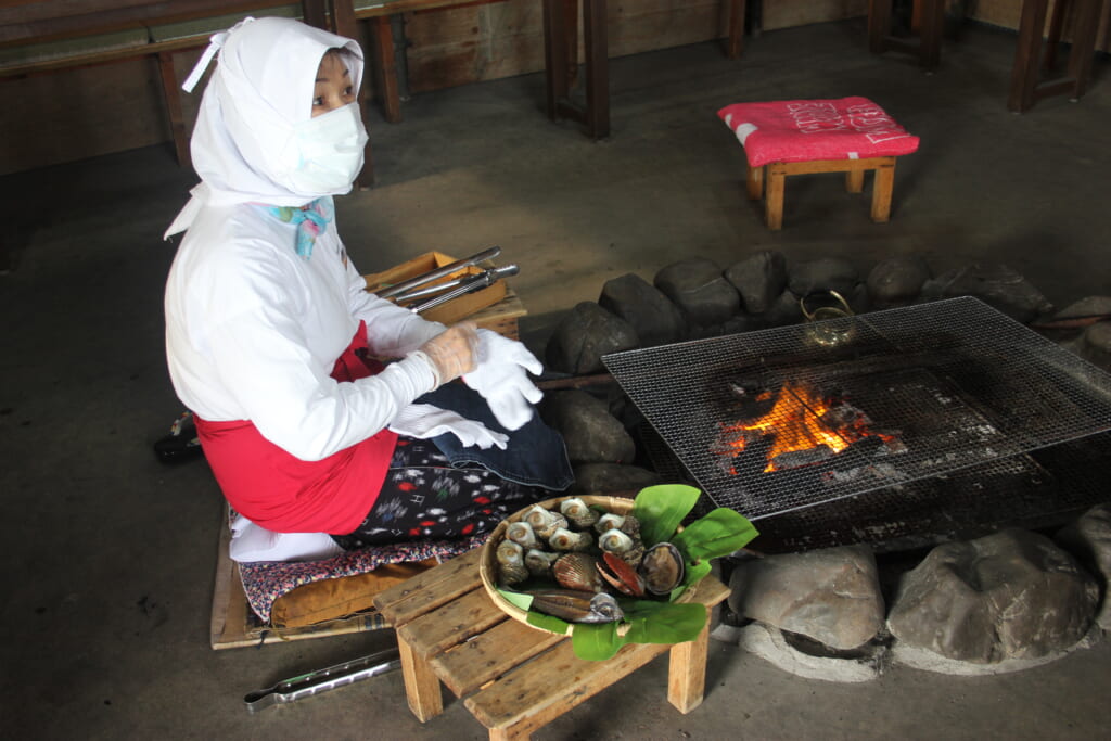 Une femme est assise sur un coussin devant un barbecue et enfile des gants. Elle s'apprête à cuisiner le plateau de fruits de mer posé à sa droite.