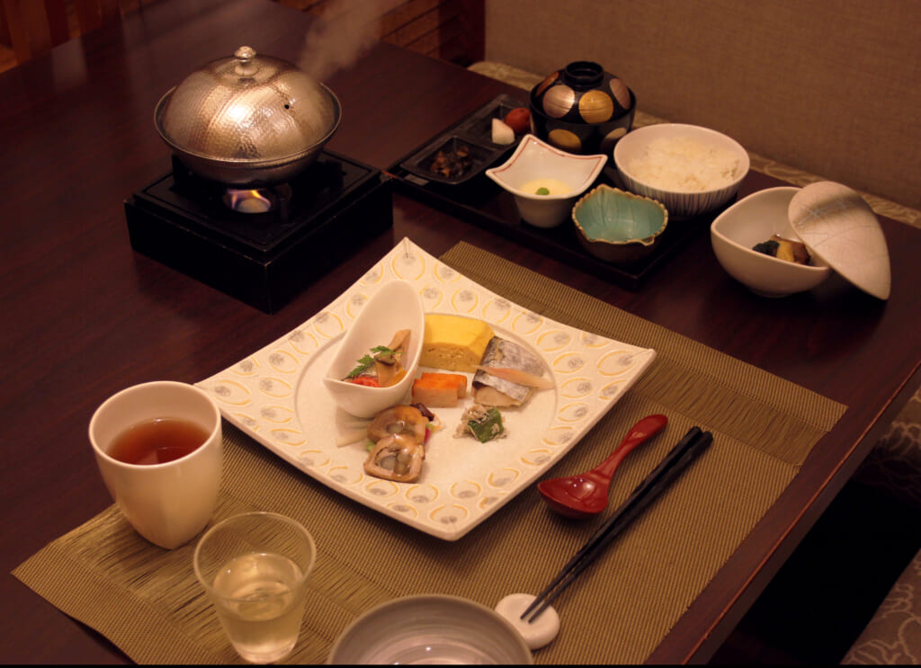 Sur une table, un petit déjeuner japonais s'apprête à être mangé. Il est composé d'une assiette principale contenant entre autre une omelette, du poisson, des rouleaux de viande. Plus loin, d'auytres assiettes plus petites sont remplies riz, de boeuf mijoté et de soupe, et une petite assiette métallique avec un couvercle fume sur un chauffe-plat.