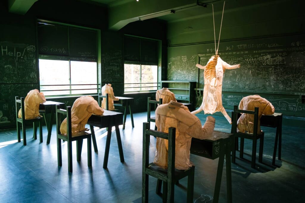 Bárbara Sánchez-Kane, Versos Rancios : installation de chemises figurant des élèves et d'une carcasse en tissus dans une salle de classe peinte en vert foncé et recouverte d'inscriptions à la craie