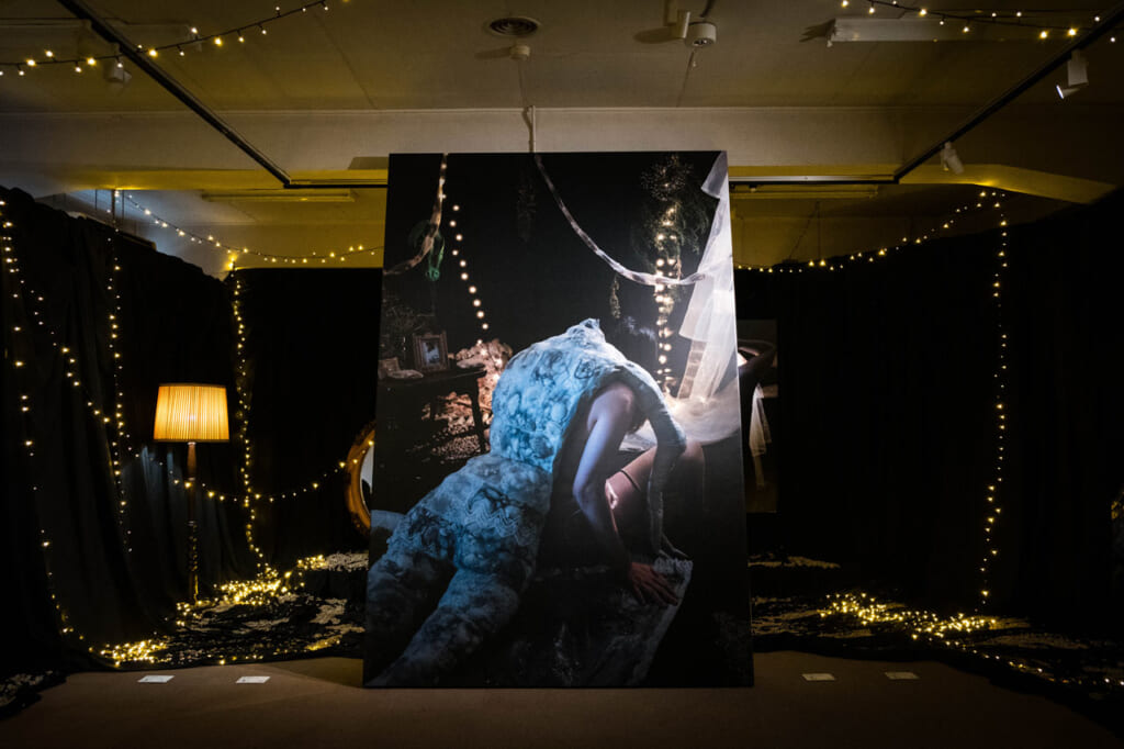 Mari Katayama, Possession : autoportrait de l'artiste avec une poupée de tissus et de coquillages sur le dos, dans une pièce décorée de tentures noires et de guirlandes d'ampoules 